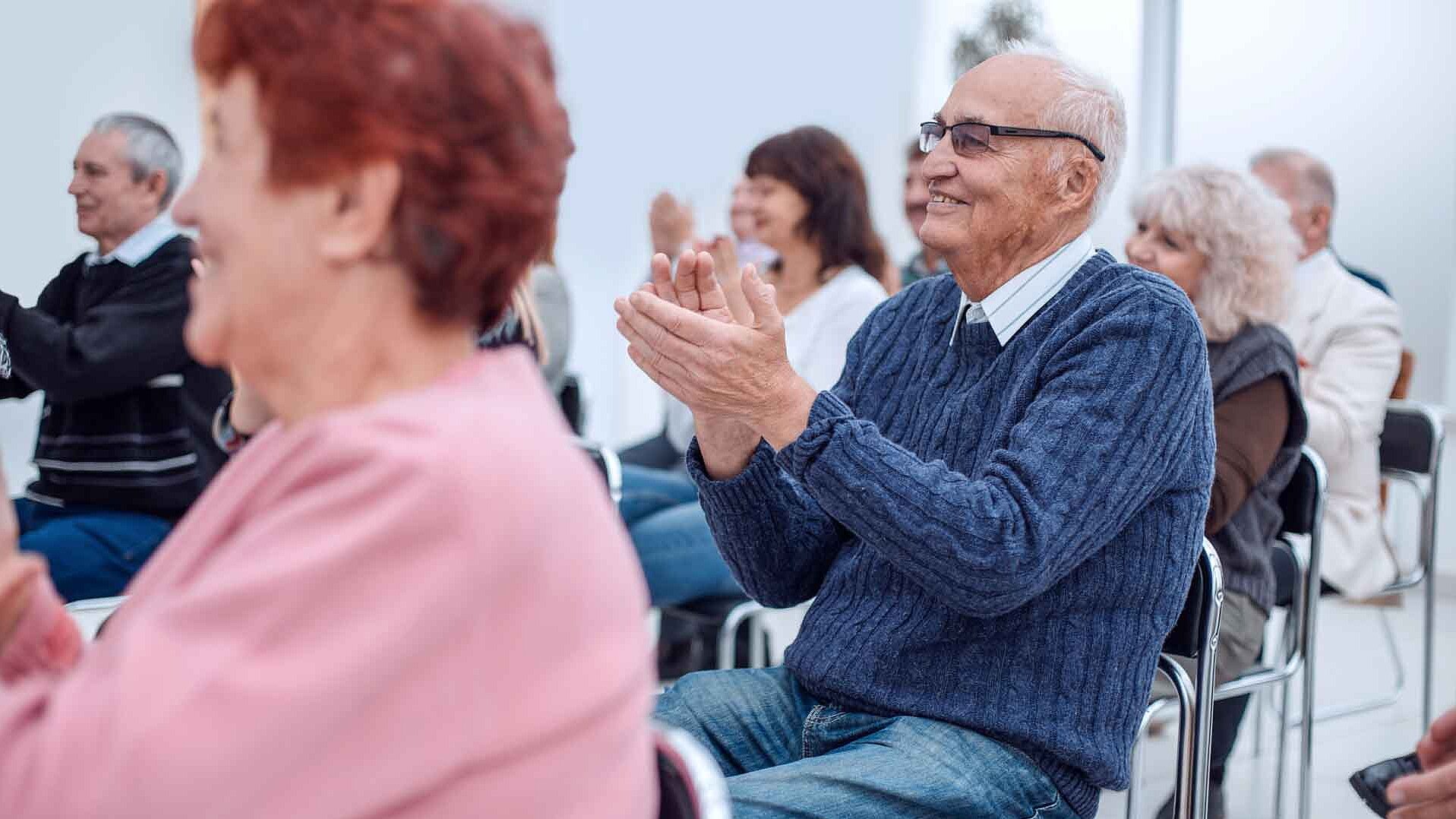 Menschen sitzen auf Stühlen in einem hellen Raum und klatschen bei einer Veranstaltung.